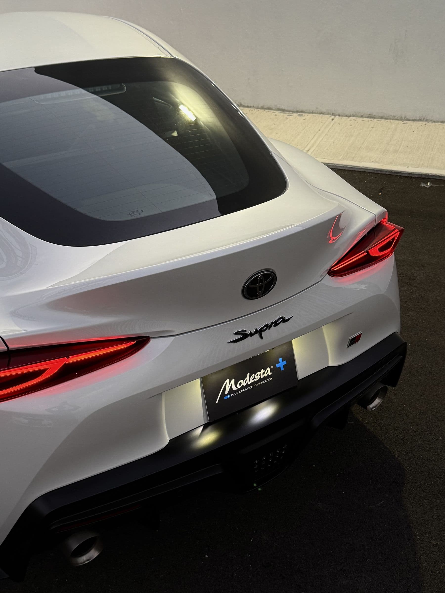Toyota GR Supra blanco, vista trasera con tablilla Modesta+ afuera del estudio